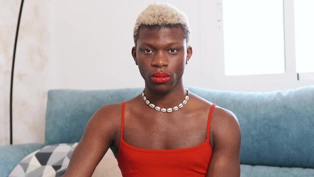 Transgender man with makeup and dress looking at camera while sitting on a sofa.