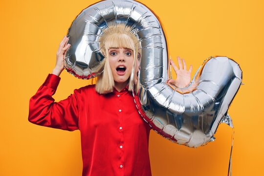 horizontal photo on a yellow background of a happy, joyful woman standing in a red shirt with a balloon in the form of the number two of silver color sticking her head into it