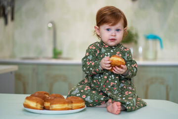 A little girl with red hair in a green dress sits on a table and eats sufganiyah donuts on Hanukkah day