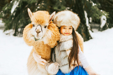 cute beautiful asian girl in fashion stylish winter clothes fur coat and fur hat walking and hugging with llama alpaca pet in snowy pine forest, winter spirit and having fun