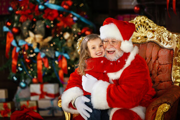 Fototapeta na wymiar Santa Claus hugs the child and promises to fulfill his cherished wish. A smiling little girl on Santa's lap.
