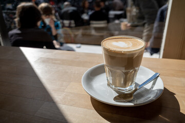 Flat white coffee in clear transparent glass and white plate lay on rough wooden table counter beside windows of coffee shop and blur background of outdoor seats and people outside coffee shop.