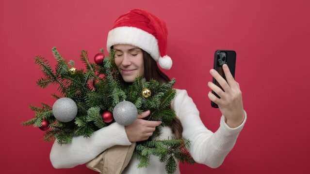 Portrait of good-looking young brunette female in Santa Claus hat holding bouquet of spruce branches do selfie shot on mobile cell phone post photo on social network, posing over red studio background