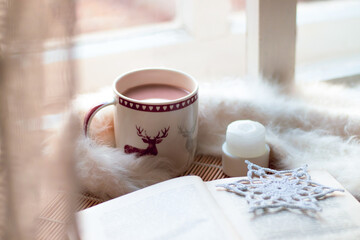 christmas still life. cup of coffee, open book, white blanket, candle, handmade snowflake on windowstill near light window, selective focus