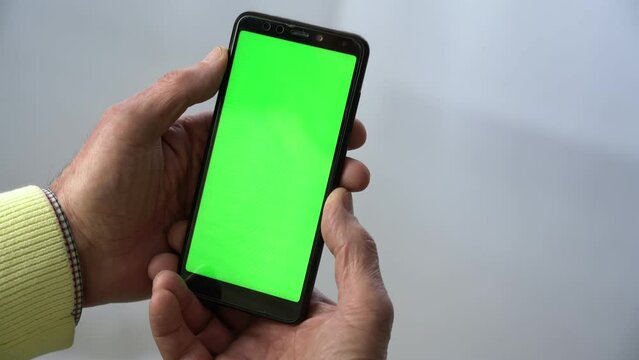 Old man holding smartphone with chromakey green screen vertically in wrinkled hands against white background at home