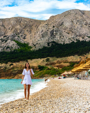 beautiful woman in a classic white dress enjoys a sunny day on a paradise beach in croatia, bay surrounded by mountains, krk island in the mediterranean sea