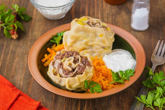 Unleavened dough rolls stuffed with minced meat and potatoes in a brown clay bowl on a wooden background. Served with fried grated carrots and sour cream.