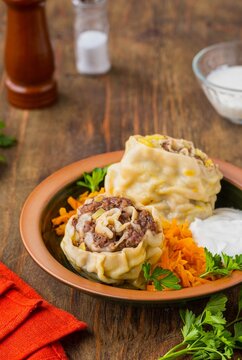 Unleavened dough rolls stuffed with minced meat and potatoes in a brown clay bowl on a wooden background. Served with fried grated carrots and sour cream.