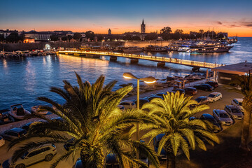 Zadar, Croatia - Aerial view of palm trees at the city of Zadar at golden sunset sky with illuminated City Bridge (Gradski most), Cathedral of St. Anastasia tower and yacht marina - obrazy, fototapety, plakaty
