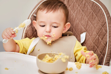 Happy toddler baby boy learns to eat porridge himself with a spoon while sitting in a child chair....