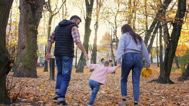 Happy family walking together in the autumn park. Mom, dad,and jumping daughter walk holding hands on autumn leaves outdoors in city park.Parenthood, carefree childhood, love and happy family concept.