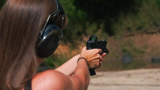 Closeup outdoor back-view of skinny caucasian girl with long dark brown hair wearing protective headphones and preparing to aim at target with loupe rifle. High quality 4k footage