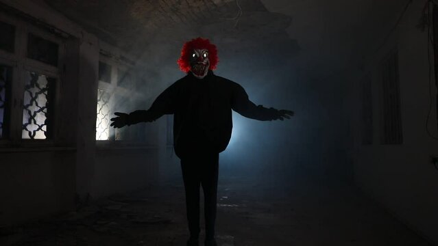 Scary evil clown with red hair standing inside ruined hall in abandoned building at night. Selective focus