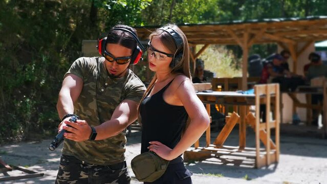 Medium outdoor shot presenting two caucasian people at shooting range. Learning long-haired woman in black tank top and body-builder-like male instructor in moro shirt. High quality 4k footage