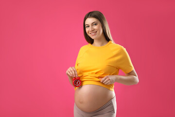 Young pregnant woman holding alarm clock near her belly on pink background. Time to give birth