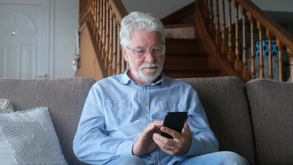 One old man using phone at home sitting on the sofa smiling and having fun learning chatting with friends and surfing on the web. Satisfied male senior indoor with smartphone.