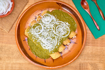 Mexican food (green mole enchiladas) served with onion rings and aged cheese in a clay dish on a wooden table, decorated with colorful napkins of the Mexican flag. top view