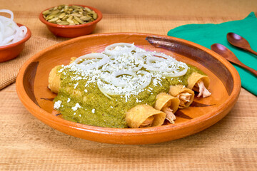 Mexican food (green mole enchiladas) served with onion rings and aged cheese in a clay dish on a wooden table, decorated with colorful napkins of the Mexican flag.close up view