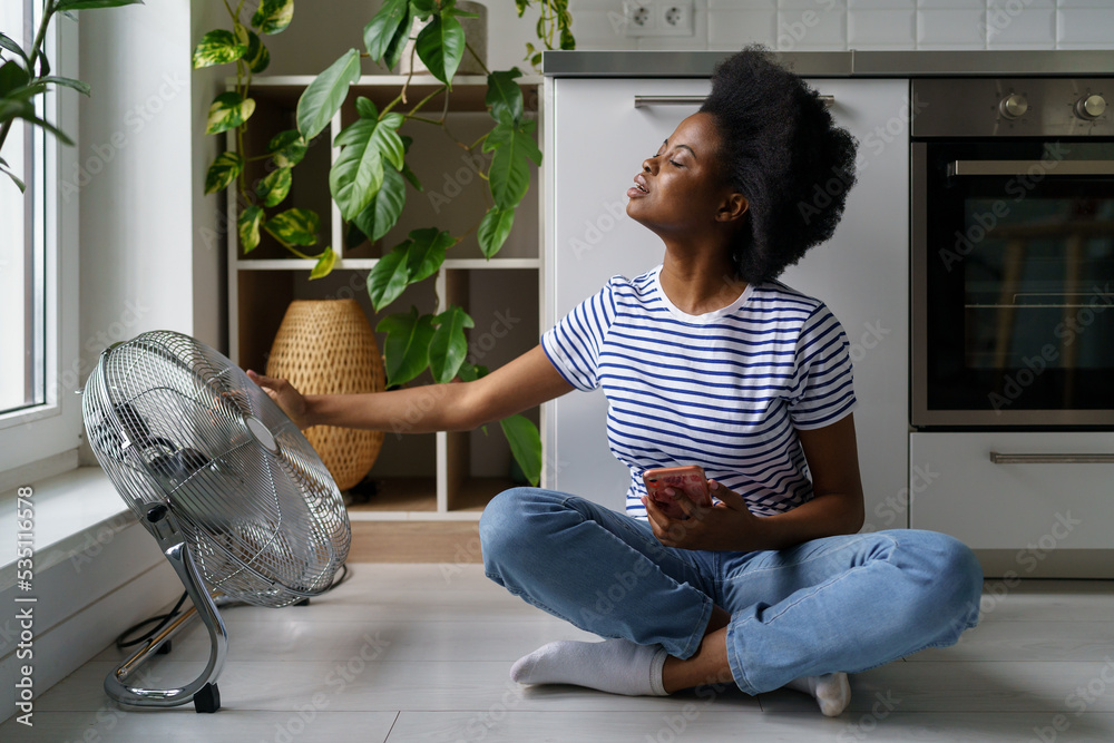 Wall mural young black woman sits on floor with smartphone, tries to catch air currents emanating from electric