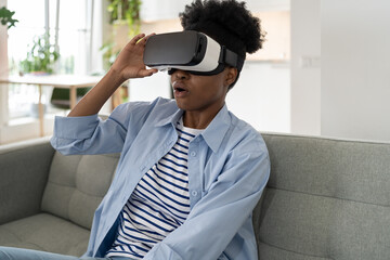 Shocked African American woman using vr glasses at home. Virtual reality and wearable tech concept with young black female wearing headset goggles. Digital technology for entertainment or leisure