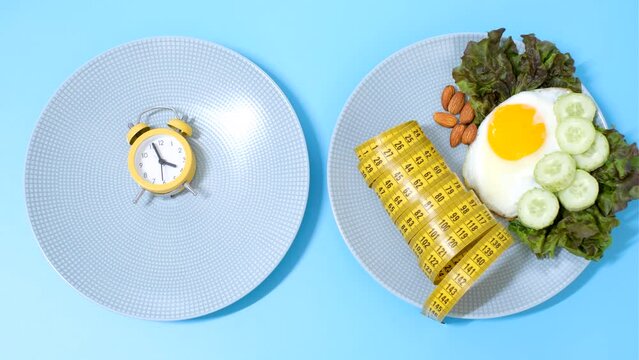 Female hand puts an alarm clock on plate of food on blue background, concept of intermittent fasting.