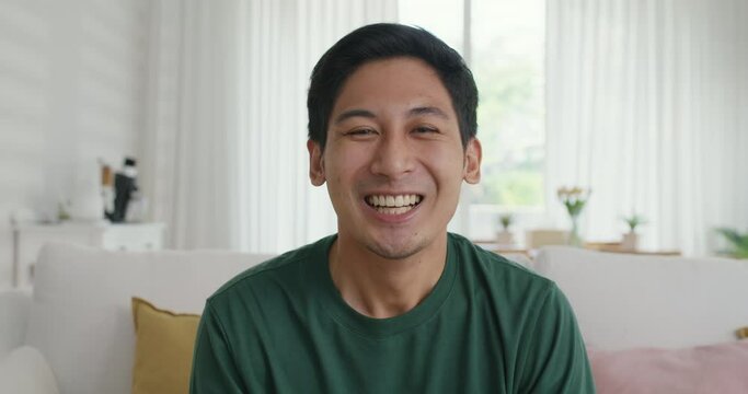 Close-up adult male Gen Z asia people relax look at webcam camera toothy smile sit at home sofa couch. New house owner young latin man guy happy joy cheerful face in urban modern health care lifestyle