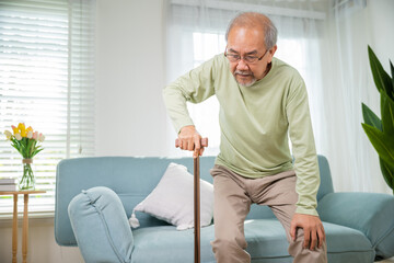 Asian senior old man with eyeglasses type to standing up from sofa with walking cane stick to walk...