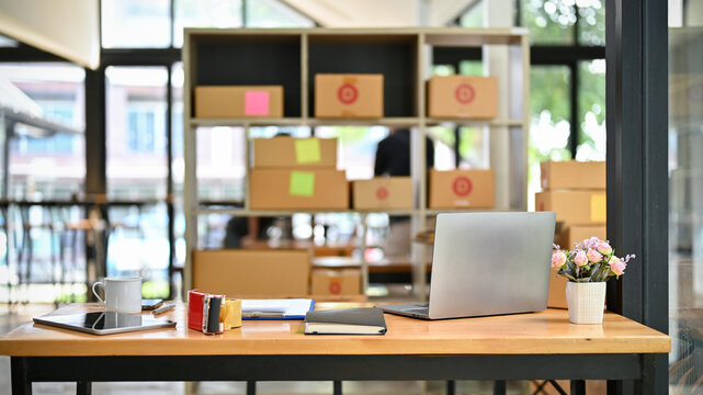 Office desk in a packing room with laptop, supplies, cardboard boxes and packing equipment.