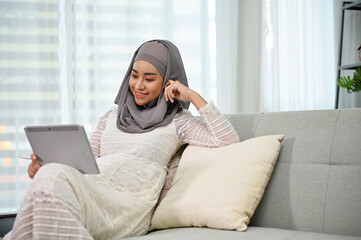 Asian Muslim pregnant woman enjoying watching video on tablet while resting in her living room.