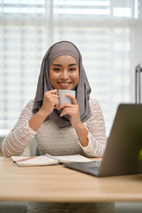 Beautiful Asian Muslim woman wearing hijab enjoying her coffee while working at her desk.