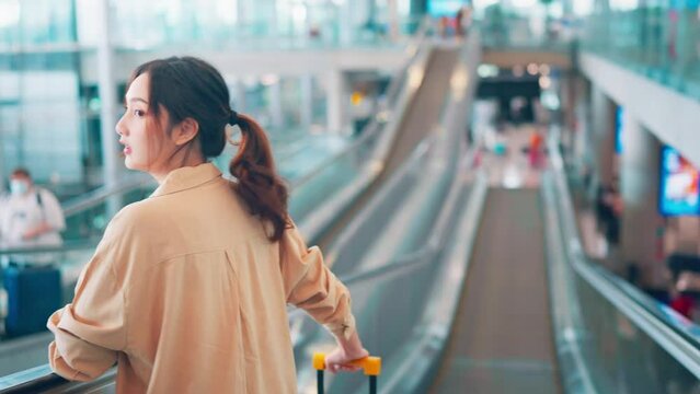 Rear view Asian tourist woman traveller with simple suitcase stand on moving walkway in airport terminal, Tourist journey trip concept
