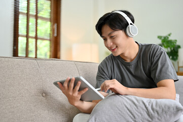 Asian man listening to music, using tablet while relaxing in living room.