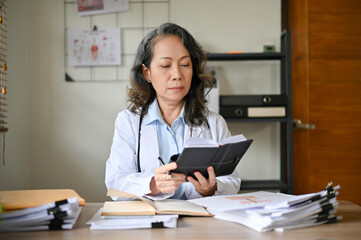 Professional senior Asian female doctor at her office desk, checking her surgery schedule.