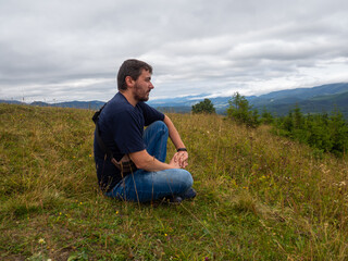 Naklejka na ściany i meble Young man admiring breathtaking view while sitting on the ground in the mountains. Man in casual clothes sits on the hill on cloudy summer day. Vacation trip to the Carpathians.