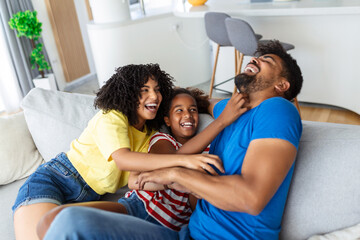 Cheerful people sitting on couch in living room have fun little daughter tickling mother laughing together with parents enjoy free time playing at home. Weekend activity happy family lifestyle concept - obrazy, fototapety, plakaty