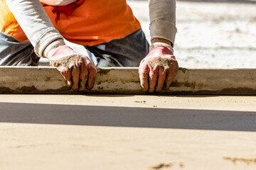 Close up of man builder placing screed rail on the floor covered with sand-cement mix at construction site. Male worker leveling surface with straight edge while screeding floor. Blurred background.