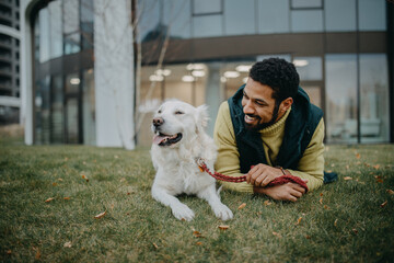 Happy young man resting with his dog outdoors in city park, during cold autumn day.