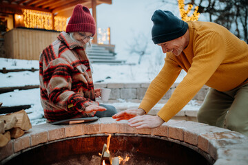 Senior couple sitting and heating together at outdoor fireplace in their winter garden.