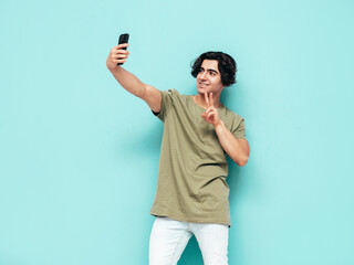 Portrait of handsome smiling stylish hipster lambersexual model.Man dressed in over size T-shirt and jeans. Fashion male isolated in studio. Posing near blue wall. Taking selfie