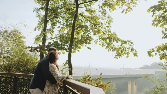 Behind view young lovely couple stay closely together near railing in autumn park, embracing, kissing, enjoy spend time together outdoors look at distance dreaming about future.