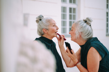 Senior women, twins in city walk, completing make-up with lipstick.