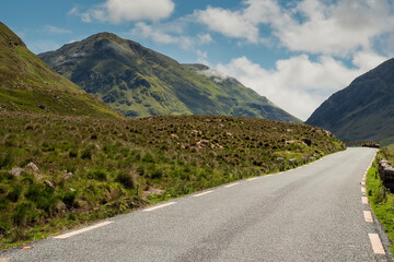 Small narrow road in a mountain area with beautiful nature scenery. Warm sunny day, cloudy sky. Connemara area, Ireland. Irish landscape.