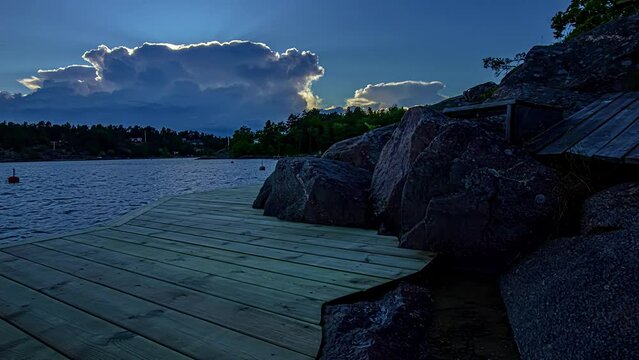 Dusk over a lake with silhouettes of dark trees in the distance. Time lapse view from the wooden pier on the shore. Buoys swaying in the waves