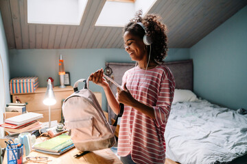 Black girl listening music while packing her backpack in bedroom