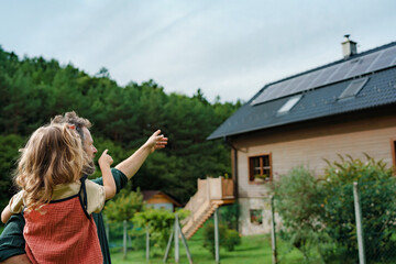 Rear view of dad holding her little girl in arms and showing at their house with installed solar...