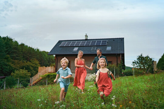 Happy family running near their house with solar panels. Alternative energy, saving resources and sustainable lifestyle concept.