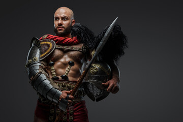 Portrait of isolated on grey background gladiator with muscular build holding helmet and spear.