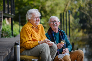 Happy senior couple having picnic and resting near lake after walk.