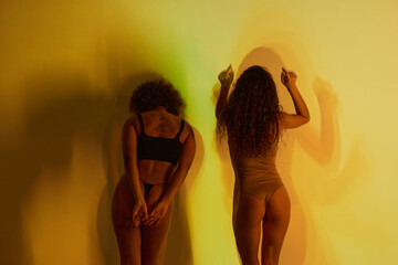 Horizontal rear view medium long studio shot of two ethnically diverse women wearing lingerie standing in yellow projector light