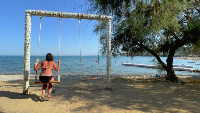 Back view of adult woman on summer holiday enjoy swinging on rope swing looking at calm sea